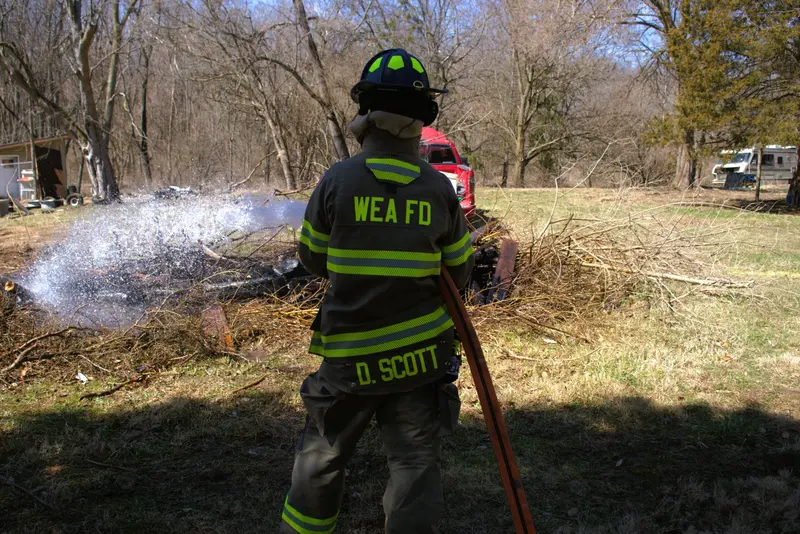 A firefighter knocks down a smoldering brush pile with a hose line in a wooded field.