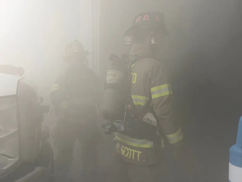 Two firefighters in full gear and air packs stand in a smoke-filled apparatus bay.