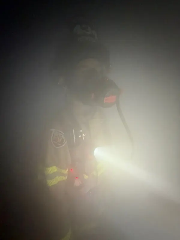 A firefighter in an SCBA mask, a chest flashlight cutting through dense training smoke.