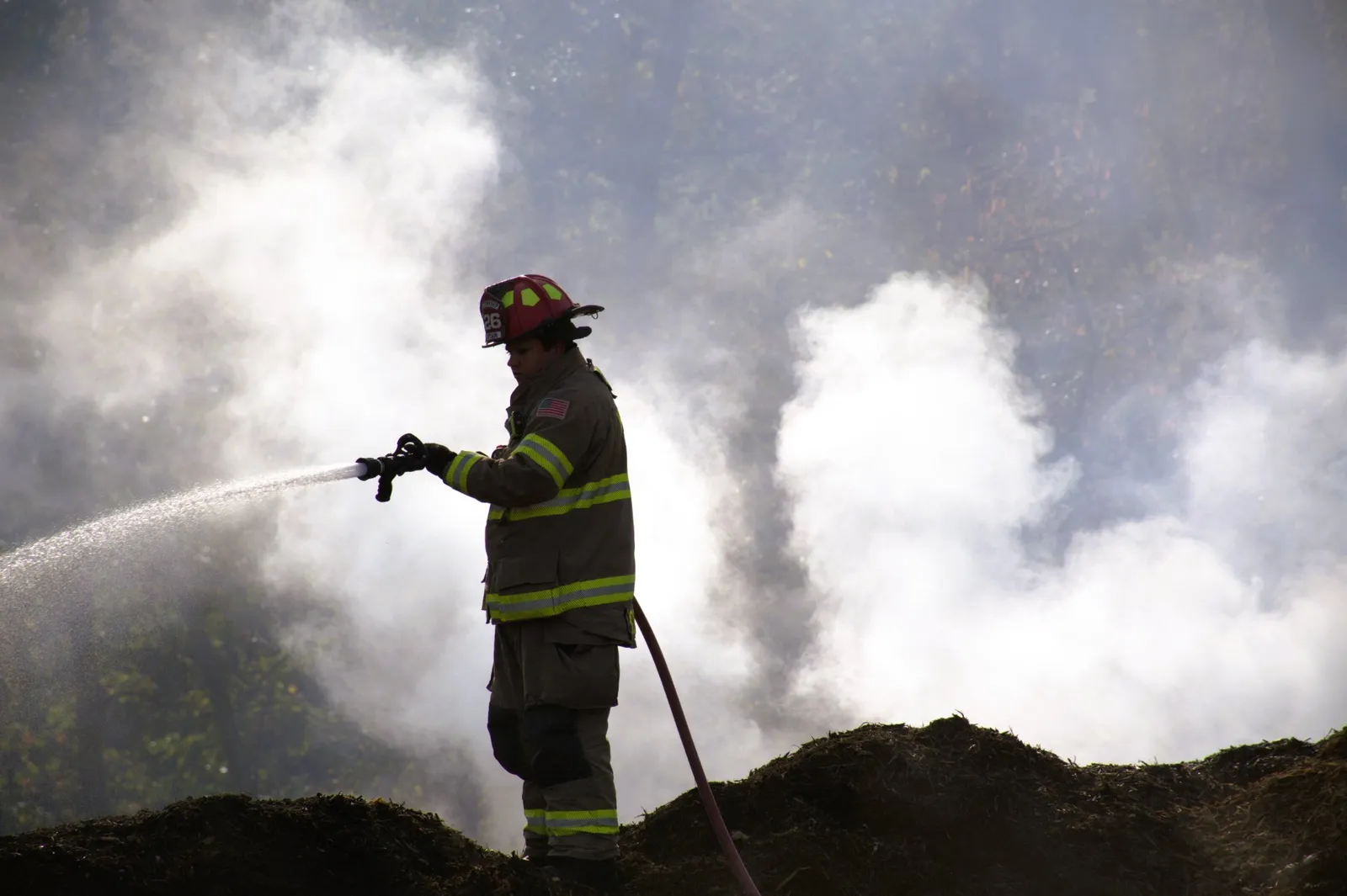 A firefighter sprays a hose line into thick white smoke rising from a mulch pile.