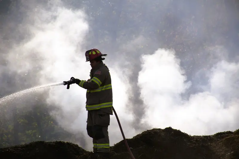 A firefighter sprays a hose line into thick white smoke rising from a mulch pile.
