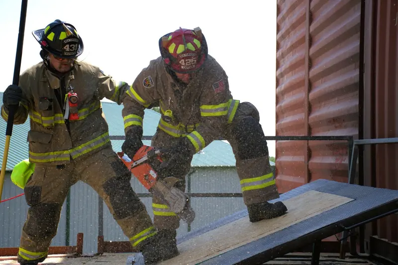 A firefighter cuts into a roof ventilation prop with a rotary saw, debris flying.